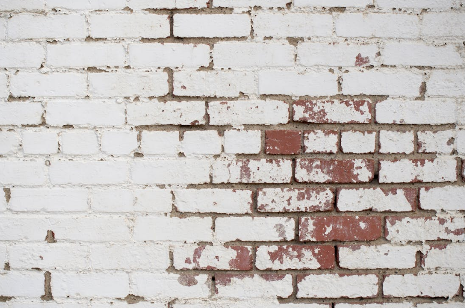 A weathered white brick wall with peeling paint revealing the original red bricks underneath.