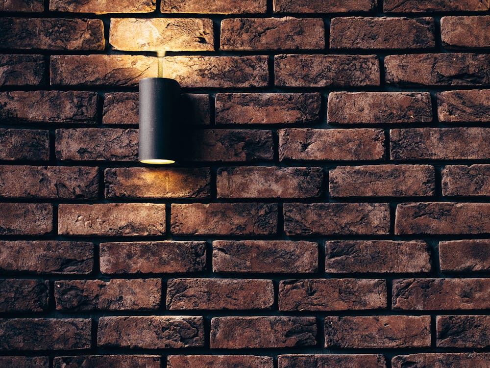 A single dark sconce casts light upwards on a textured brick wall.
