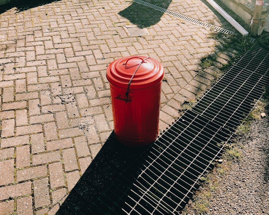 a drainage grill alongside a brick pavement and red dustbin