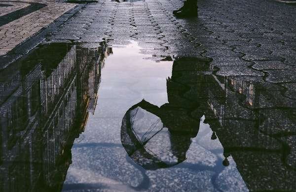  A person holding a transparent umbrella is reflected in a puddle on a rainy city street.