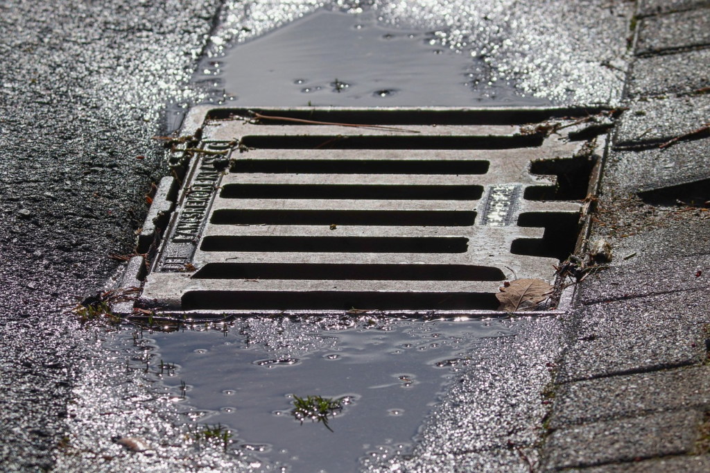 A metal storm drain grate is surrounded by wet pavement and small puddles.