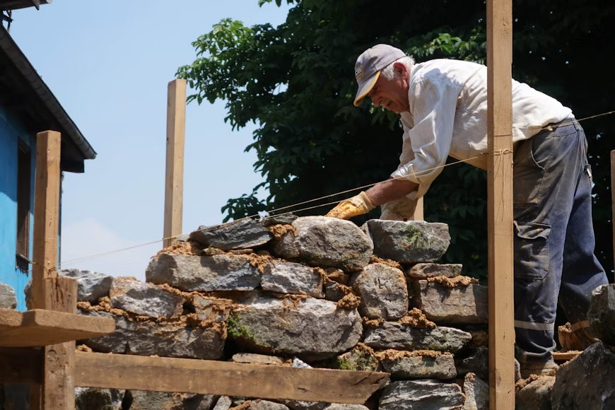 A man building a stone wall, carefully aligning rocks with the help of guide strings on a sunny day.