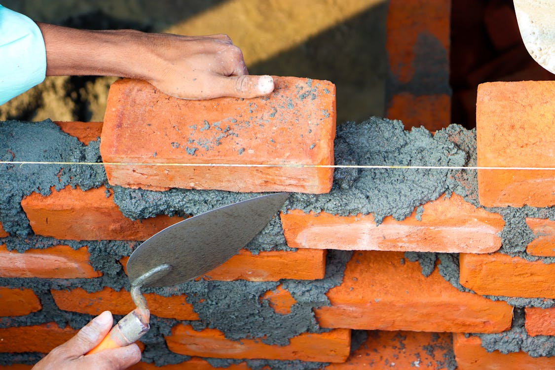 A close-up of a person laying bricks with mortar using a trowel, with a guide string for alignment.