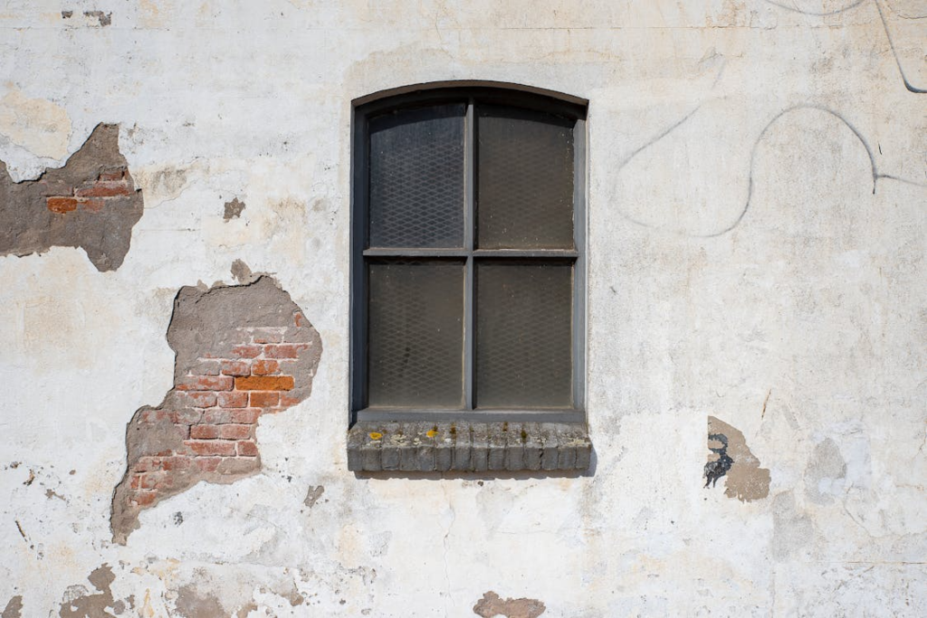 The image shows a weathered wall with peeling plaster and exposed bricks around an old window.