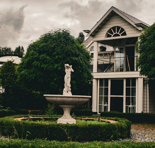  A peaceful garden featuring a stone fountain and a house with large windows, under an overcast sky.
